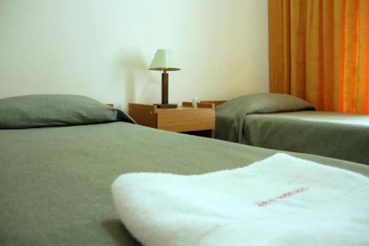 Habitación de hotel con dos camas, una mesita de noche, una lámpara y una ventana con cortinas.