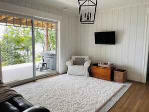 A room with a white chair, a flat-screen TV, a wooden side table, and a large window with a view of greenery.