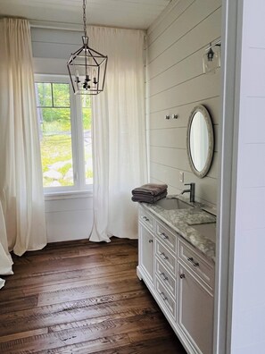 A bathroom with a wooden floor, a white vanity with a marble countertop, a hanging light fixture, and a window with white curtains.