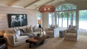 Un vaste salon doté d’une grande fenêtre donnant sur un lac, d’un plafond rustique et d’un lustre moderne.