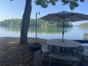 A wooden deck with picnic tables and an umbrella, overlooking a calm lake with trees in the background. - Perfect lake property for Clemson Game Day! (Seneca)