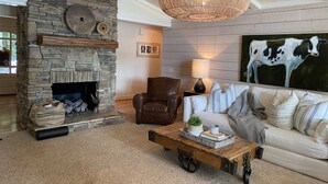 Un salon chaleureux avec une cheminée en pierre, un grand luminaire en fibres tressées, un canapé confortable et un tableau représentant une vache.