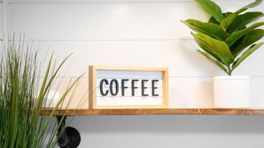 A framed sign with the word "COFFEE" on a shelf against a white wall, accompanied by a potted plant. - DT TC Suite I Queen Bed I Walk Bars + Shops (Traverse City)