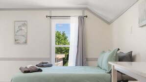 A bedroom with a bed, a small table, a window with curtains, and a view of greenery outside. - DT TC Suite I Queen Bed I Walk Bars + Shops (Traverse City)