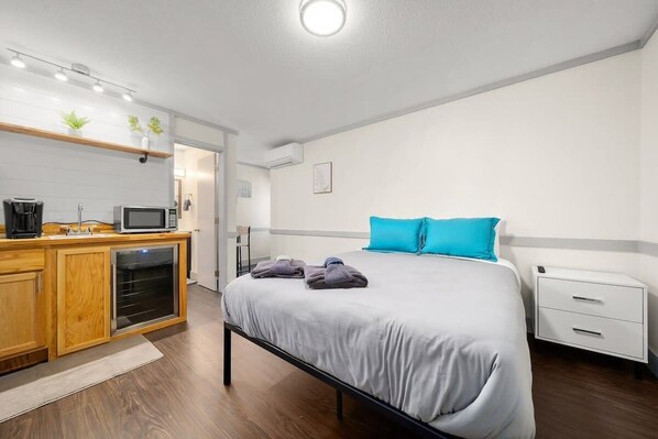 A modern hotel room with a bed, a nightstand, a desk with a computer, and a kitchenette. - Front St Suite I Queen Bed I Walk DT TC + Park (Traverse City)