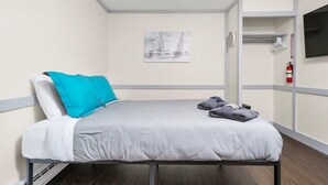Una cama bien hecha con sábana gris y almohadas azules, un pequeño cuadro de barcos de vela en la pared, y un extintor colgado en la pared.