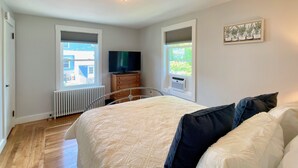 Un dormitorio con cama, cómoda, televisor y dos ventanas con persianas.