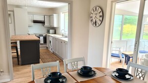 A dining area with a wooden table set for four, a kitchen in the background, and a clock on the wall.