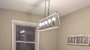A dining area with a rectangular table, four chairs, a chandelier, a window with blinds, and a 'GATHER' sign on the wall. - Peak Comfort of Apex (Apex)