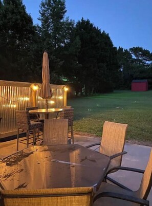 An outdoor patio with a glass table and wicker chairs, surrounded by a wooden fence with string lights.