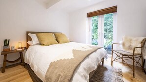 A bedroom with a bed, a chair, a nightstand, and a window with a view of greenery. - Beautiful Home in Heart of Truro (Truro)