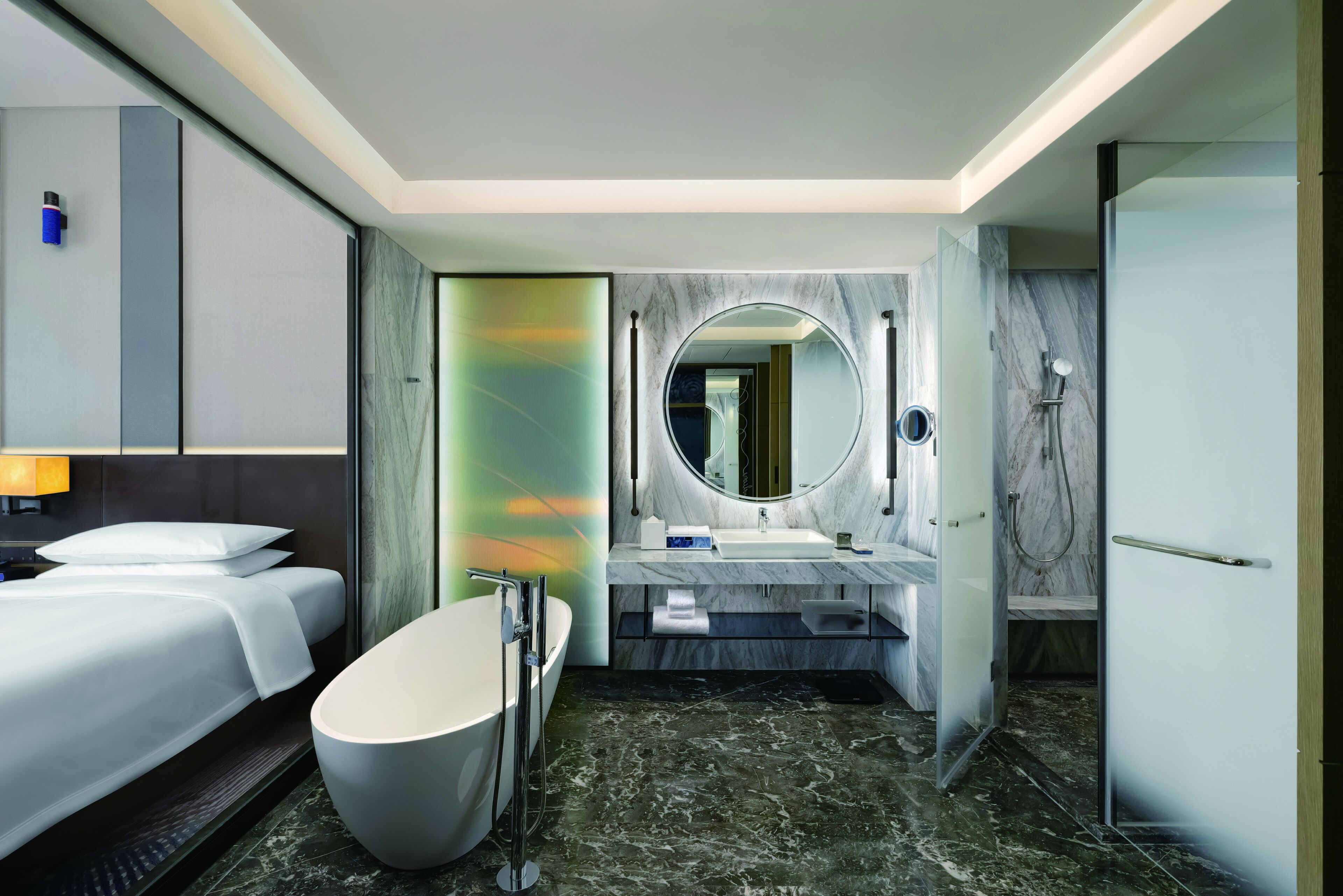 A modern hotel bathroom with a large bathtub, a round mirror, a shower area, and a separate sink.