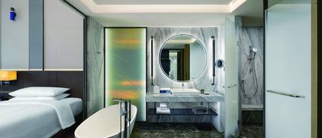 A modern hotel bathroom with a large bathtub, a round mirror, a shower area, and a separate sink.