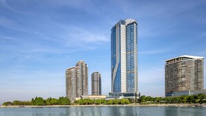A modern high-rise building with a glass facade, located near a body of water with a park and trees in the foreground. - Renaissance Changzhou Wujin Hotel (Changzhou)