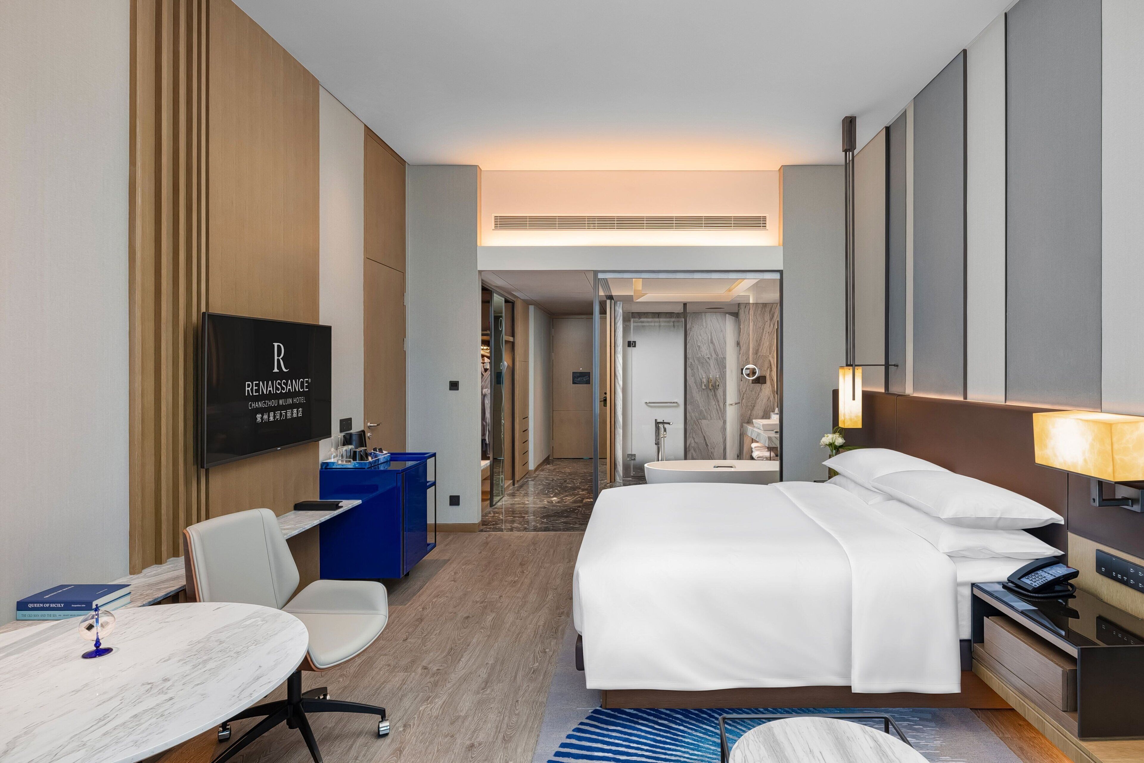 A hotel room with a bed, desk, chair, TV, and a bathroom visible in the background.