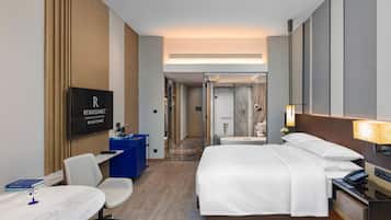 A hotel room with a bed, desk, chair, TV, and a bathroom visible in the background.