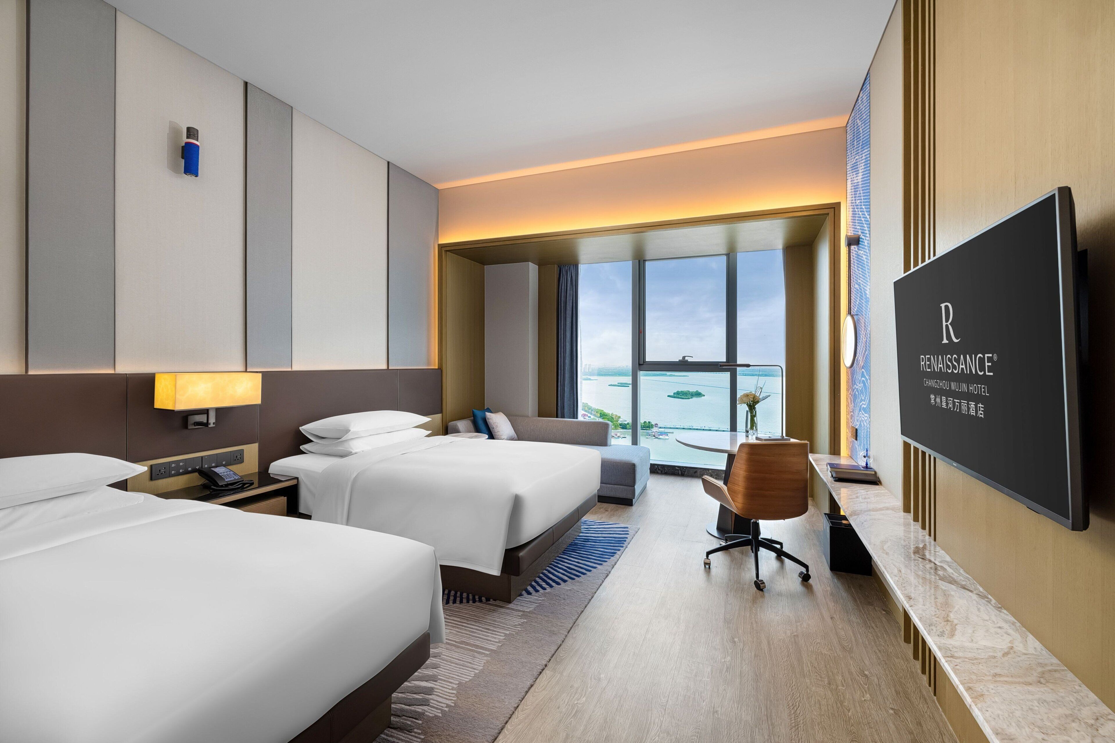 A hotel room with two beds, a desk, a chair, a television, and a large window with a view of the water.