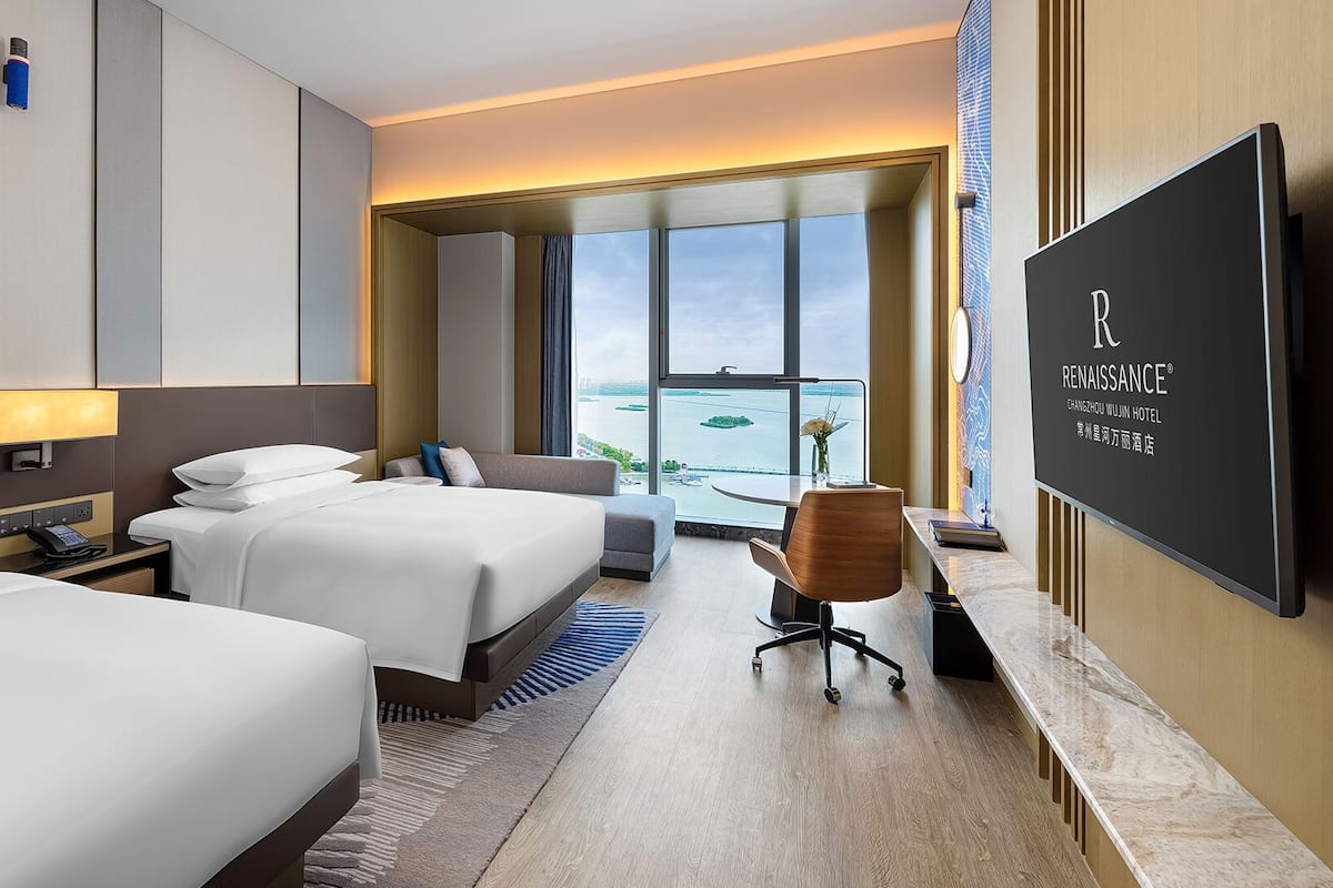 A hotel room with two beds, a desk, a chair, a TV, and a large window with a view of the water.