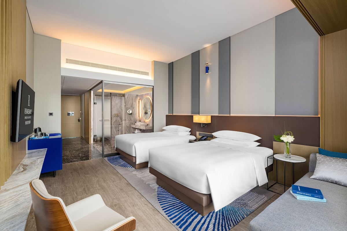 A hotel room with two beds, a desk, a chair, a small table with a vase of flowers, and a television.