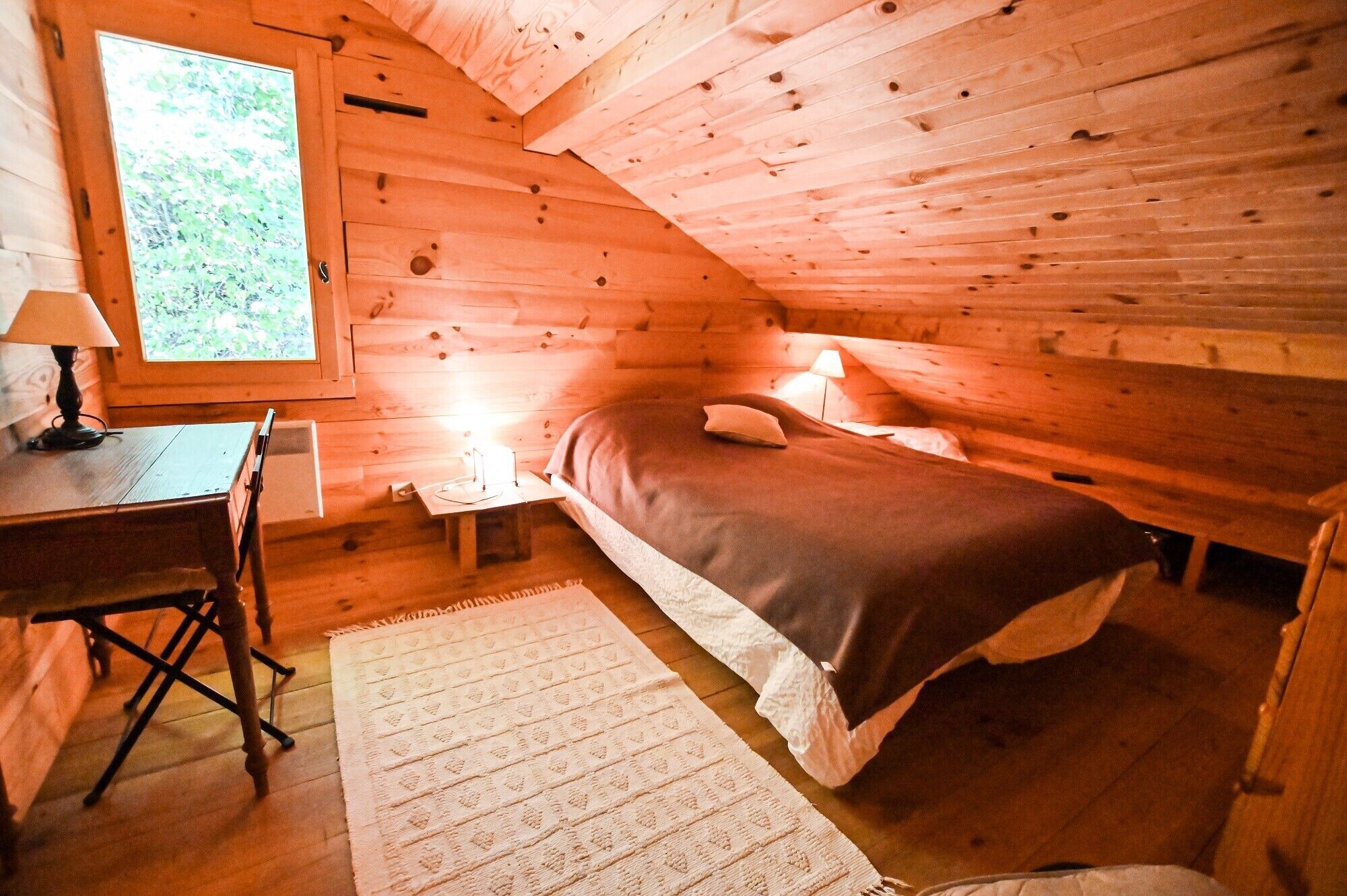 Acogedora habitación de cabaña de madera con una cama, un escritorio y una ventana con vistas a follaje.