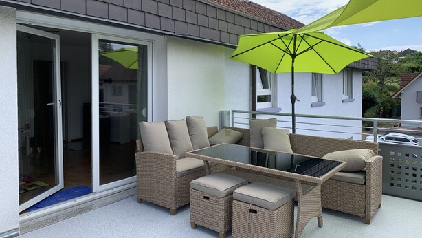 Une terrasse extérieure avec des meubles en osier, une table en verre et un parasol vert vif.