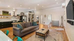 Un amplio espacio de sala con un sofá, reposapiés y una mesa de comedor. Se ve una cocina con electrodomésticos modernos y un salpicadero de baldosas tipo espiga al fondo.