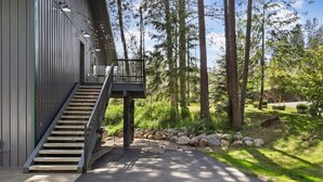 Un edificio moderno con una escalera que lleva a un balcón, rodeado de una zona boscosa con un muro de piedra y un camino pavimentado.