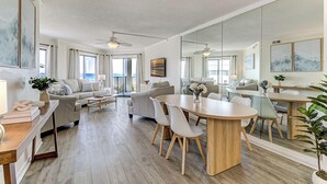 A spacious living area with a dining table, sofas, and a large mirror reflecting the room. - Oceanfront Balcony Views, Pool,  STEPS to Beach, Restaurants, Shops (Wrightsville Beach)
