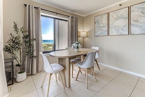 Comedor con una mesa redonda, cuatro sillas, una planta en maceta y vista al océano a través de ventanas amplias.