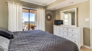 A bedroom with a large bed, a dresser, a television, and a balcony with curtains. - Ocean Views, Balcony, Pool,  STEPS to Beach Restaurants Shops (Wrightsville Beach)