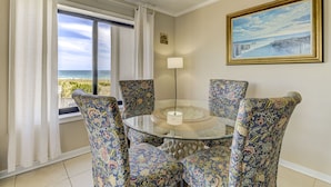 A dining area with a glass table surrounded by floral-patterned chairs, a view of the ocean through a window, and a framed painting on the wall. - Ocean Views, Balcony, Pool,  STEPS to Beach Restaurants Shops (Wrightsville Beach)