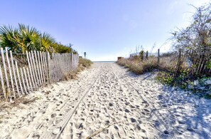 Un sentier sablonneux menant à une plage, bordé d’une clôture en bois et de végétation.