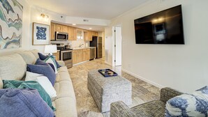 A living room with a sofa, ottoman, and a flat-screen TV mounted on the wall. - Ocean Views, Balcony, Pool,  STEPS to Beach Restaurants Shops (Wrightsville Beach)