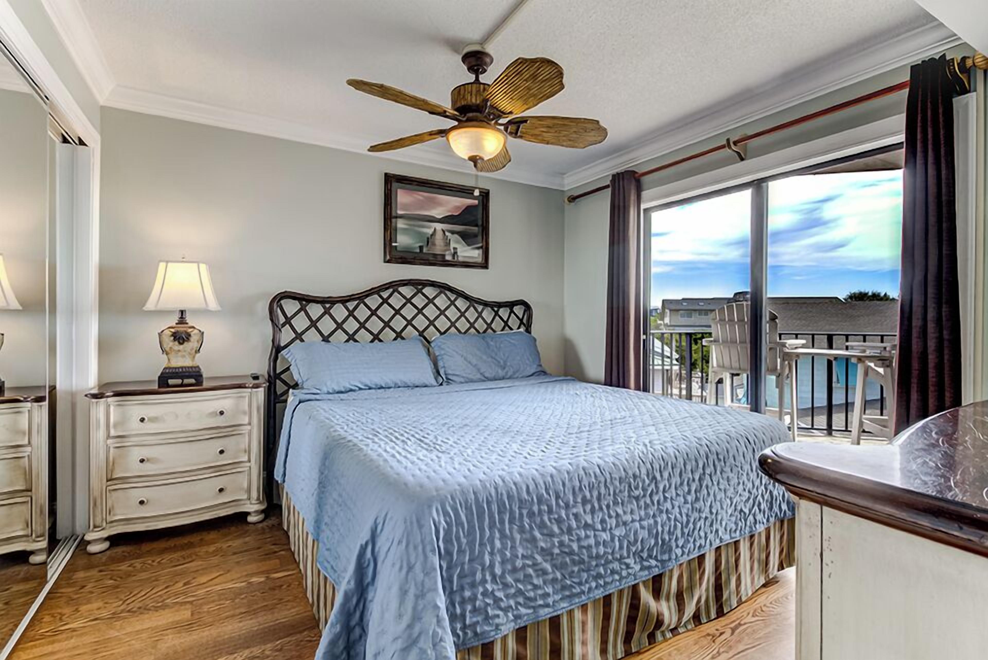 A bedroom with a bed, a nightstand, a lamp, a ceiling fan, a window with curtains, and a framed picture on the wall.