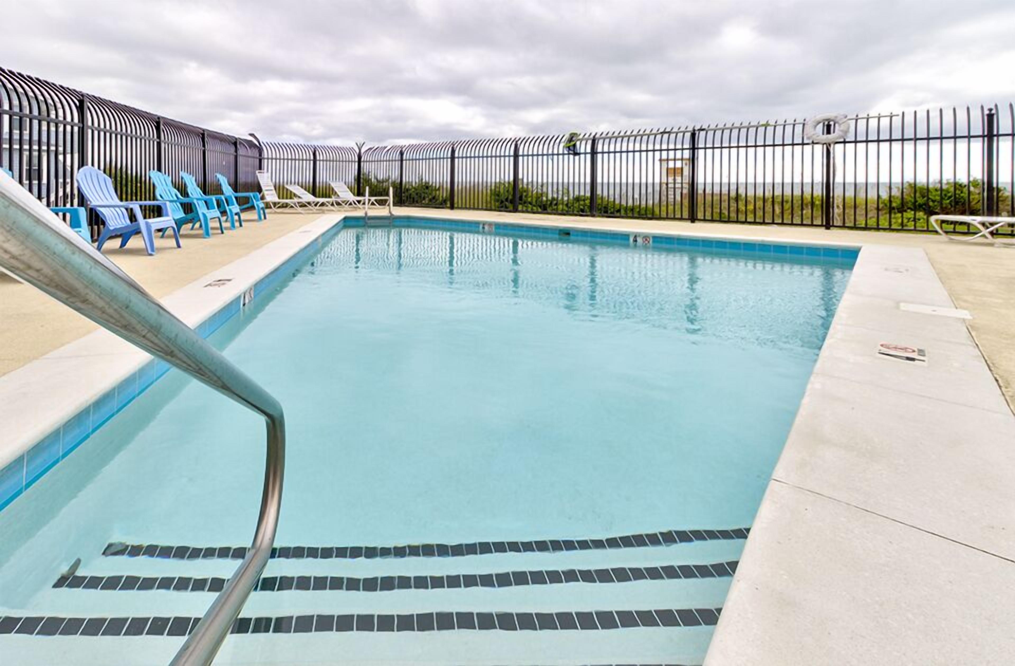 Une piscine rectangulaire avec un escalier en métal sur le côté gauche, entourée d’une clôture en métal noir. Plusieurs chaises longues bleues sont disposées le long de la clôture.