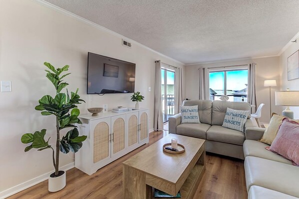 A living room with a sofa, a coffee table, a TV mounted on the wall, and a potted plant. - Ocean Views, Balcony, Pool, STEPS to Beach, Restaurants, Shops (Wrightsville Beach)