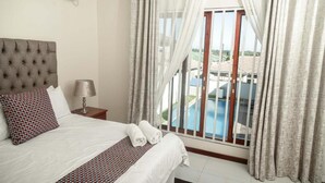 A hotel room with a bed, a nightstand, a lamp, and a view of a swimming pool. - Grayville Apartments #2 (Lusaka)
