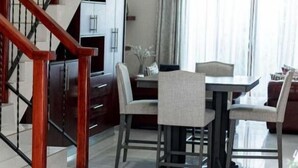 A modern living room with a wooden staircase, a dining area with a table and chairs, and a kitchen in the background. - Grayville Apartments #2 (Lusaka)