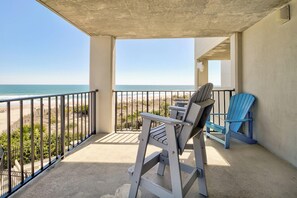 A balcony with a view of the beach, a table, two chairs, and a blue Adirondack chair.