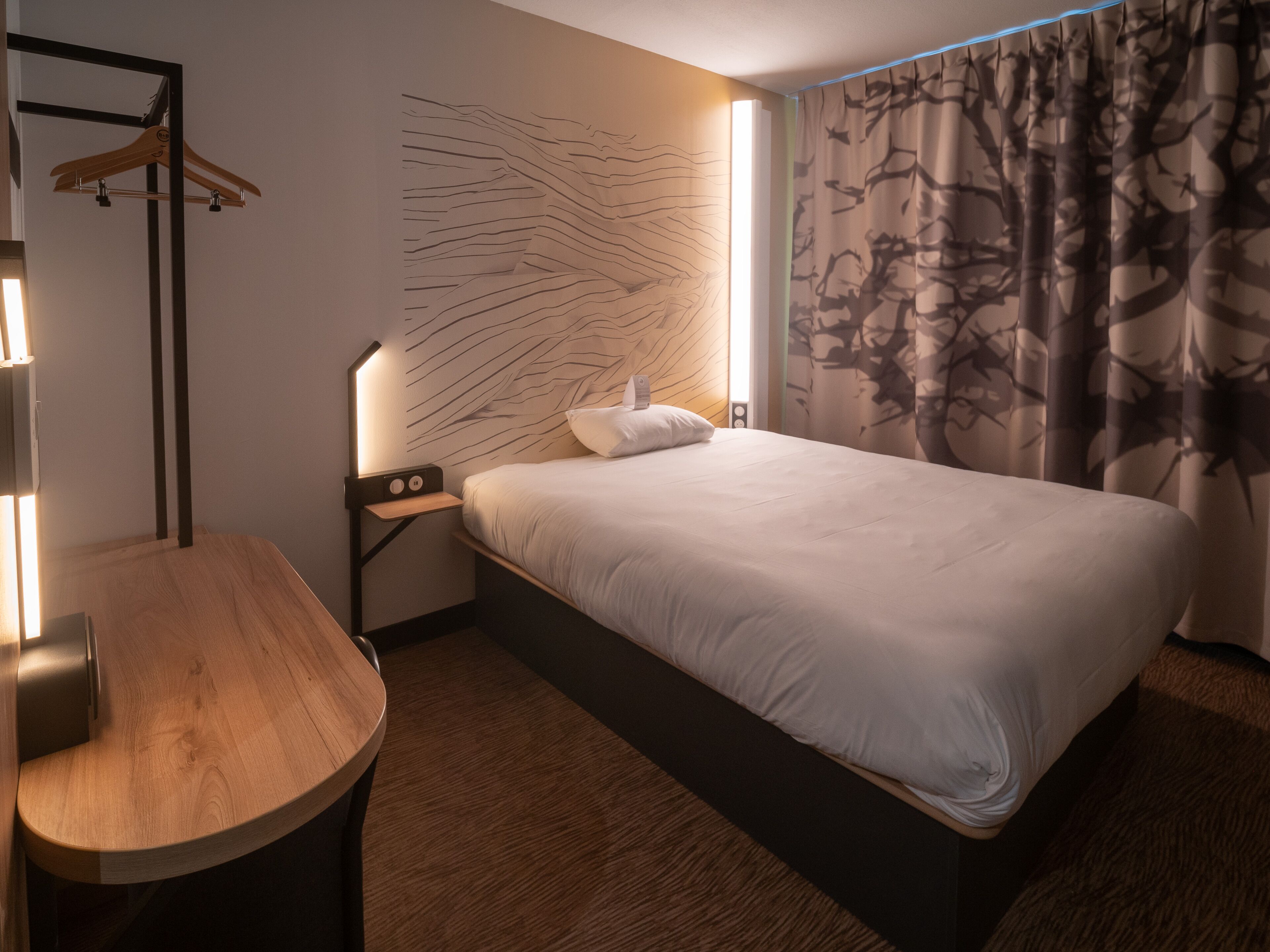 Una habitación de hotel moderna con una cama bien hecha, una mesita de noche de madera con una lámpara y una pared con un diseño abstracto único.