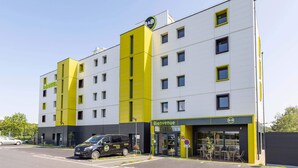 A modern hotel with a yellow and white facade, a visible entrance, and a parking area with a black van. - B&B Hotel Rennes Parc Expo Aéroport (Bruz)