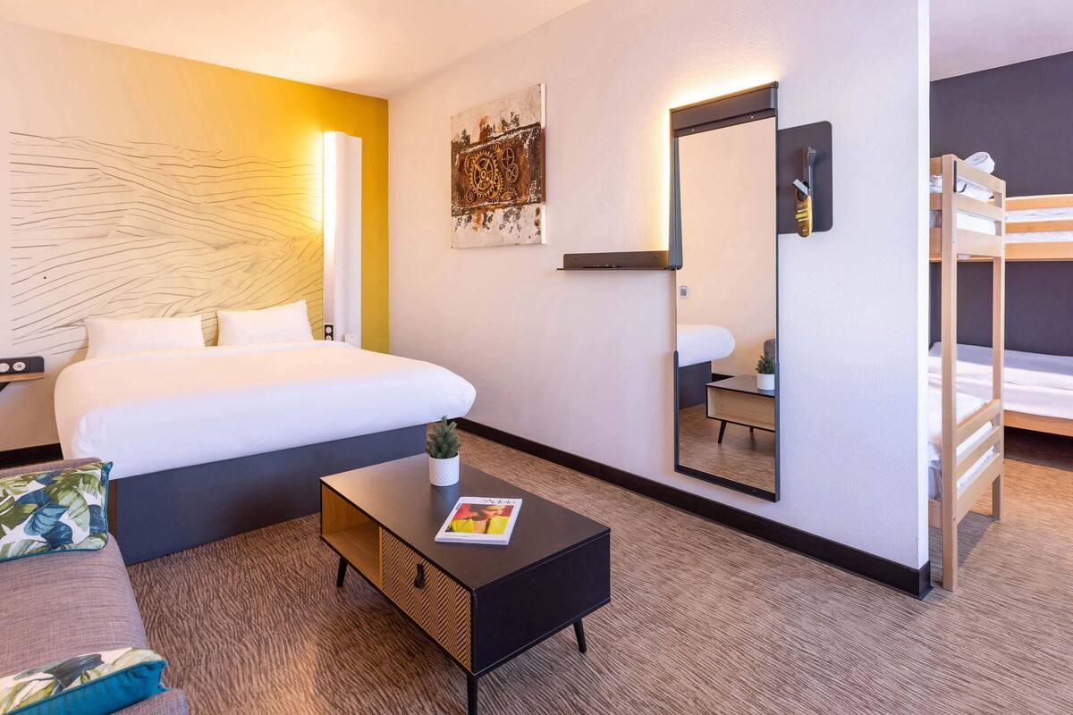 A hotel room with a bed, a small side table, a mirror, and a bunk bed.