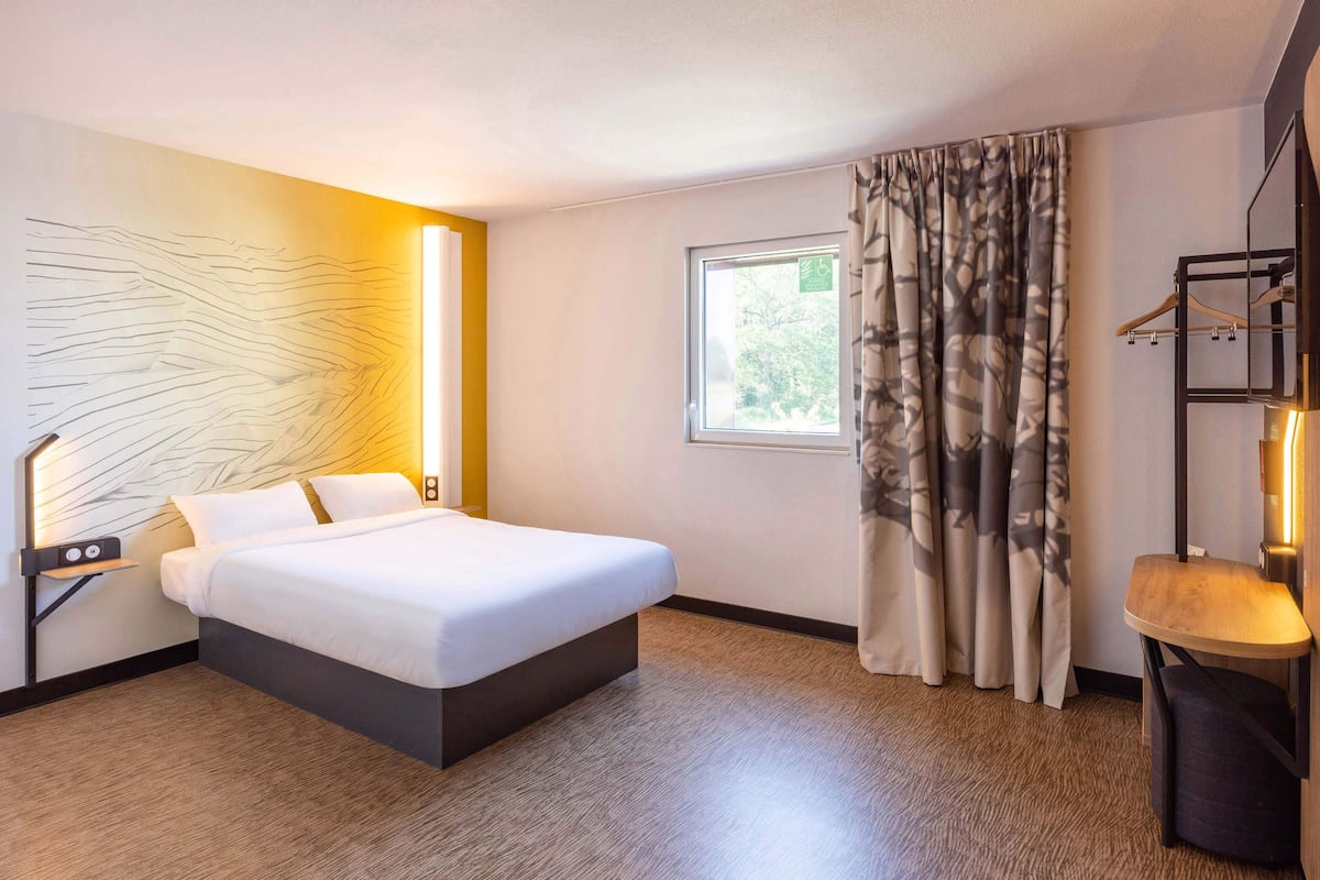 A hotel room with a bed, a bedside table, a wall-mounted light, a window with curtains, and a view of trees outside.