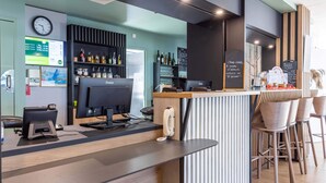 A modern bar area with a wooden counter, bar stools, and a computer monitor. There are shelves with bottles and glasses, and a clock on the wall. - B&B Hotel Rennes Parc Expo Aéroport (Bruz)