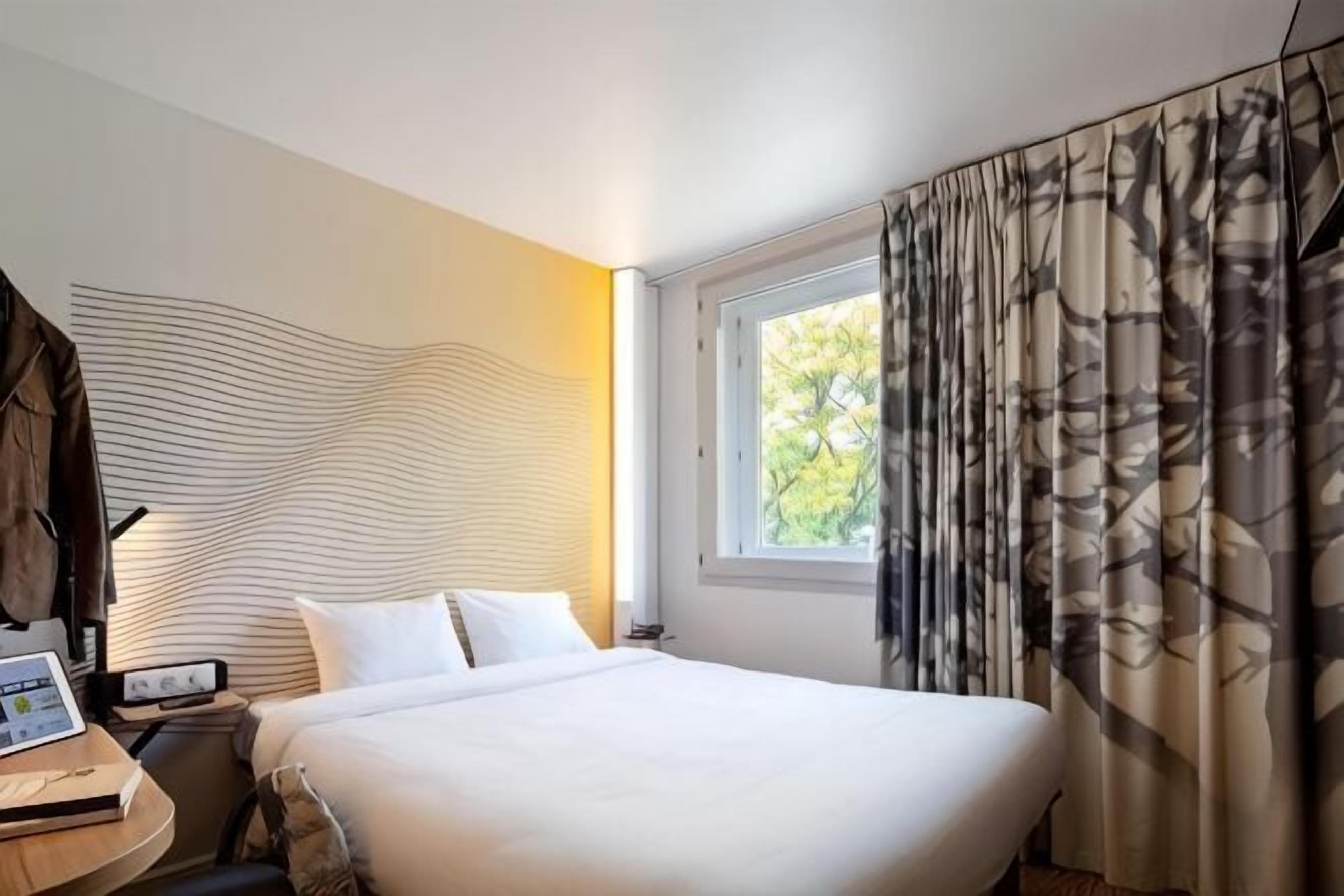 A hotel room with a bed, a desk with a laptop, a nightstand, and a window with curtains.