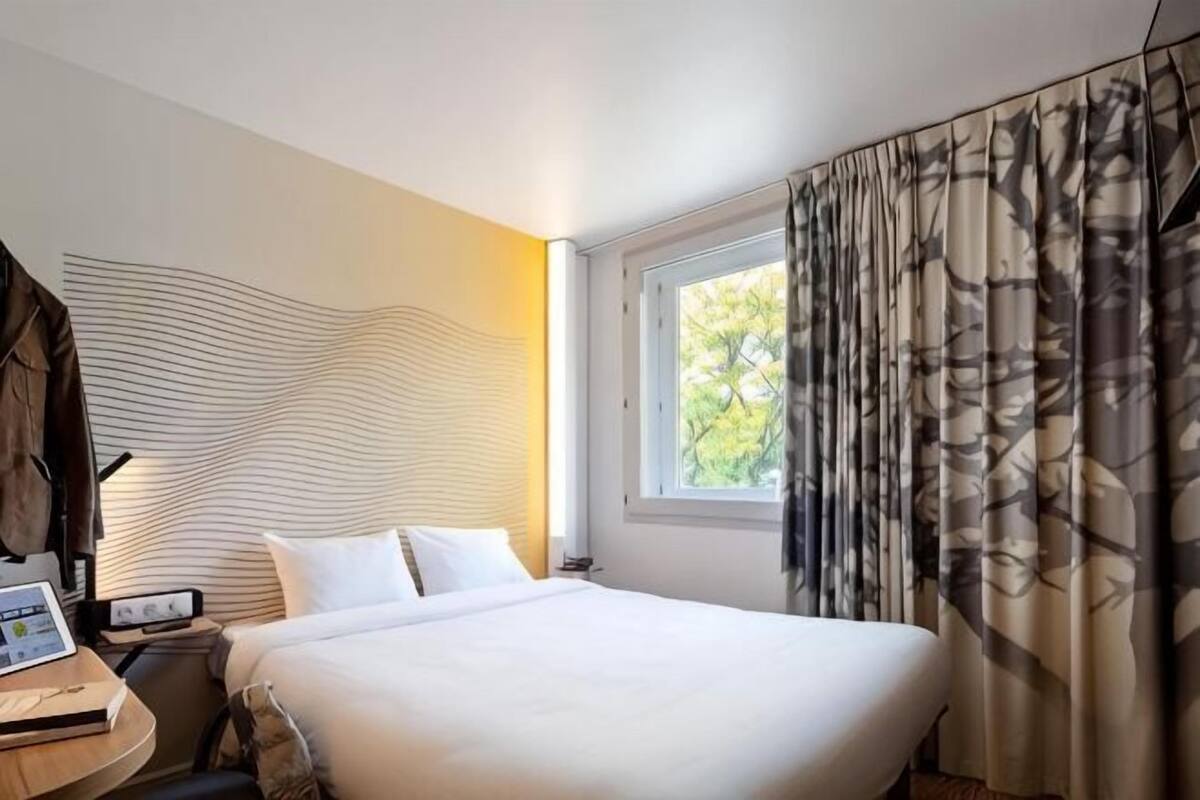 A hotel room with a bed, a desk with a laptop, a nightstand, and a window with curtains.