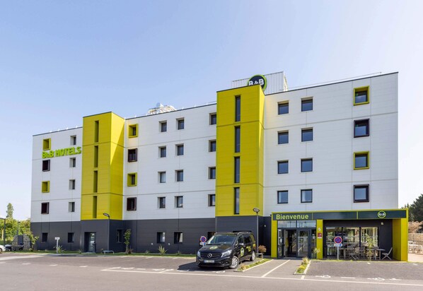 Ein modernes Hotelgebäude mit einer markanten Fassade in Gelb und Weiß, einem sichtbaren Eingang mit der Aufschrift "Bienvenue" und einem Parkplatz mit einem schwarzen Auto.