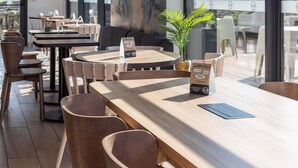 A modern cafe with wooden tables and chairs, a menu stand, and a large window with a view of the outdoors. - B&B Hotel Rennes Parc Expo Aéroport (Bruz)