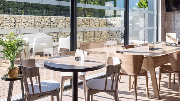 An outdoor seating area with round tables and chairs, a stone wall, and a view of trees and a fence. - B&B Hotel Rennes Parc Expo Aéroport (Bruz)