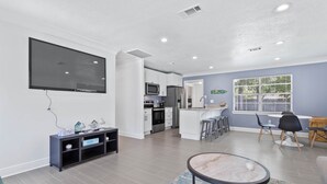 A modern living room with a flat-screen TV, a dining area with a bar, and a kitchen with stainless steel appliances.